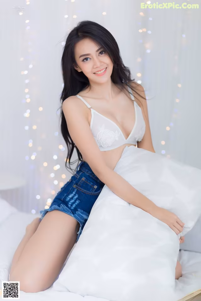 A woman sitting on top of a bed in a white bra and denim shorts.