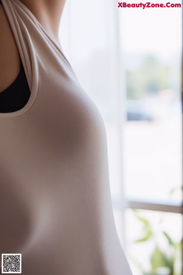 A woman wearing a white tank top standing by a window.