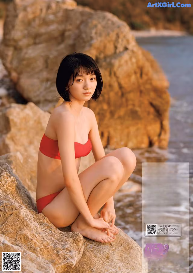 A woman in a red bikini sitting on a rock by the water.
